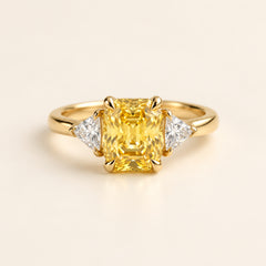 Gold ring with a yellow gemstone and two smaller diamonds on a textured surface with green leaves in the background.