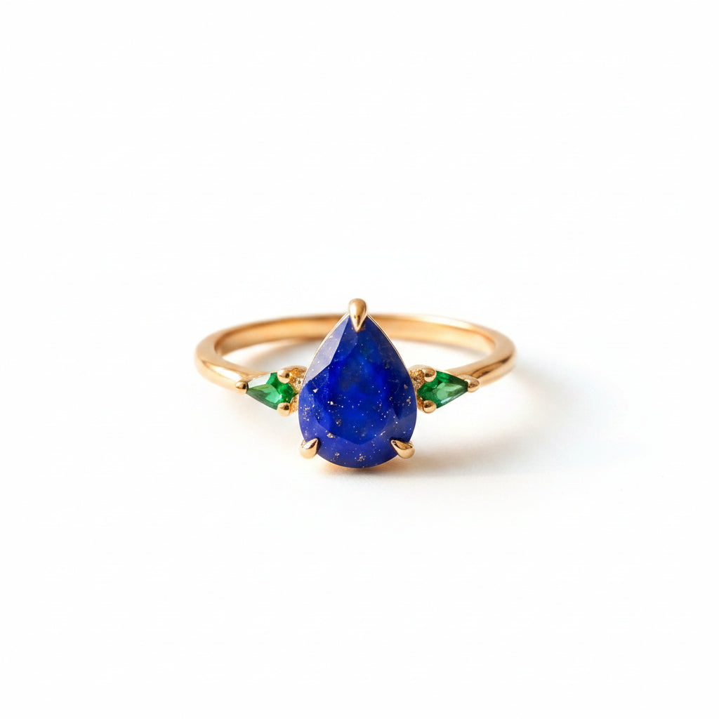 Gold ring with a blue teardrop gemstone and green accents on a textured surface with flowers.