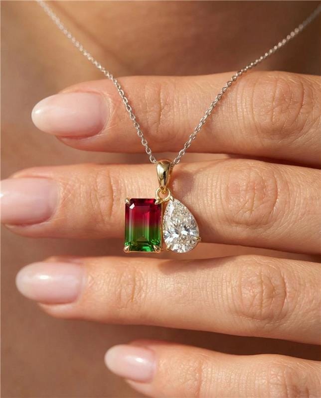 Gold necklace with a green and red gemstone pendant held by a hand.