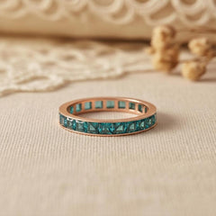 May include: Rose gold ring featuring a band of square, teal gemstones. The ring is displayed on a neutral surface, with a blurred background. The ring is a piece of jewellery.