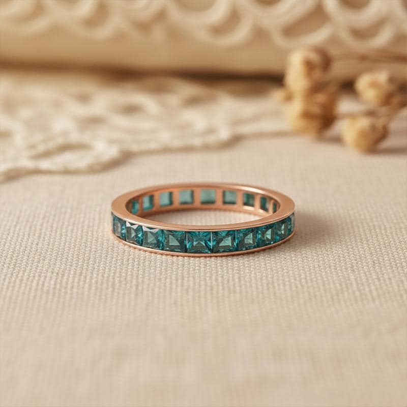 May include: Rose gold ring featuring a band of square, teal gemstones. The ring is displayed on a neutral surface, with a blurred background. The ring is a piece of jewellery.