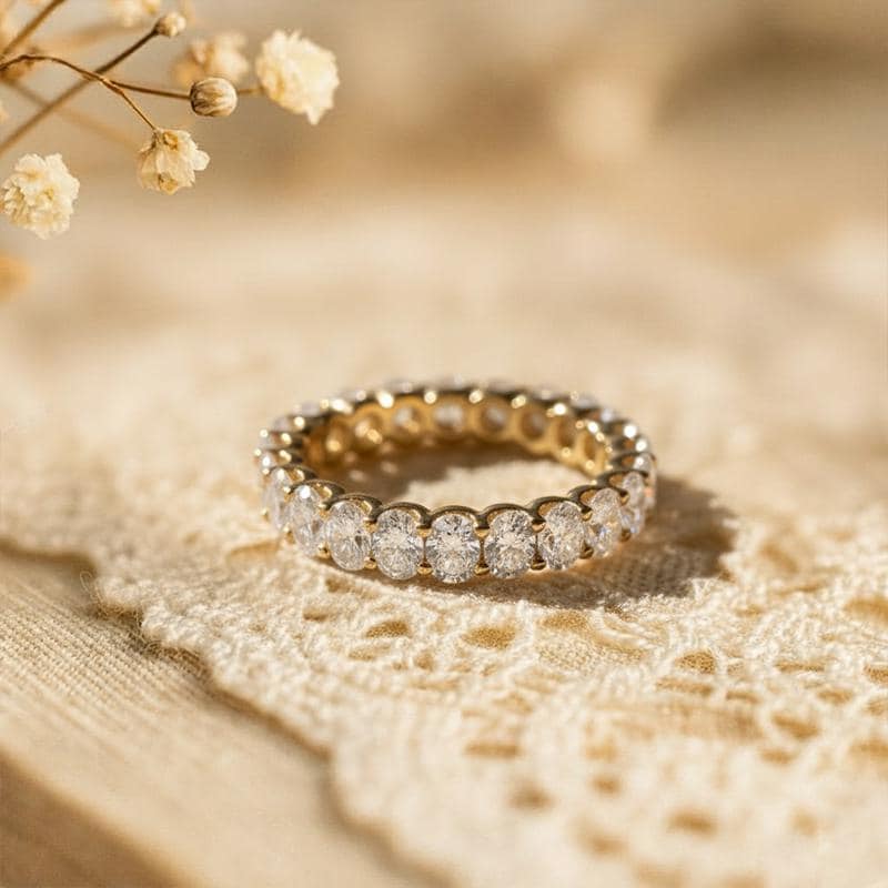 May include: A gold eternity ring featuring oval-cut clear gemstones. The ring is set on a cream lace surface, with soft lighting and a blurred background. The ring is a jewelry item.