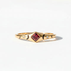 A gold ring with a central red gemstone and small side details, resting on a rustic wooden surface under warm light.
