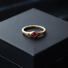 A gold ring with a central red gemstone, displayed on a dark gray box.