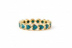 A gold band ring featuring a row of heart-shaped turquoise inlays, resting on a rustic wooden surface with a warm, softly blurred background.