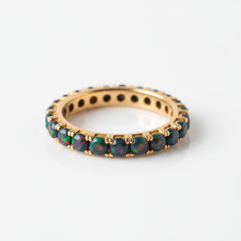A gold eternity ring featuring round black opal gemstones that shimmer with vibrant multicolor flashes, displayed on a soft, draped cream fabric.
