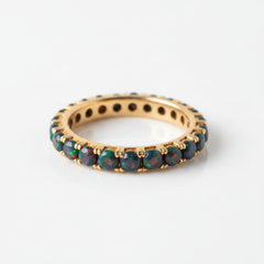 A gold eternity ring featuring round black opal gemstones that shimmer with vibrant multicolor flashes, displayed on a soft, draped cream fabric.