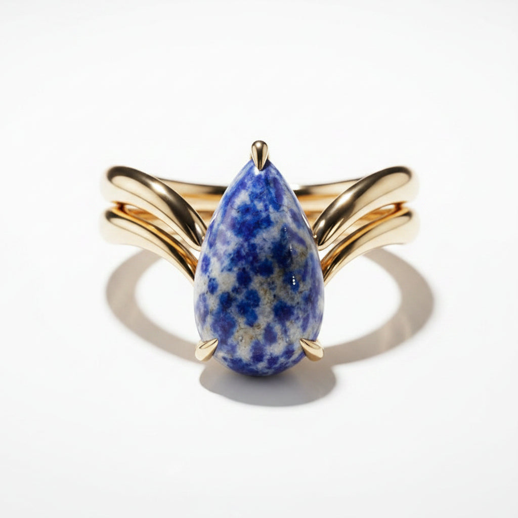 A gold ring featuring a teardrop-shaped blue and white spotted stone, displayed on a black marble surface.