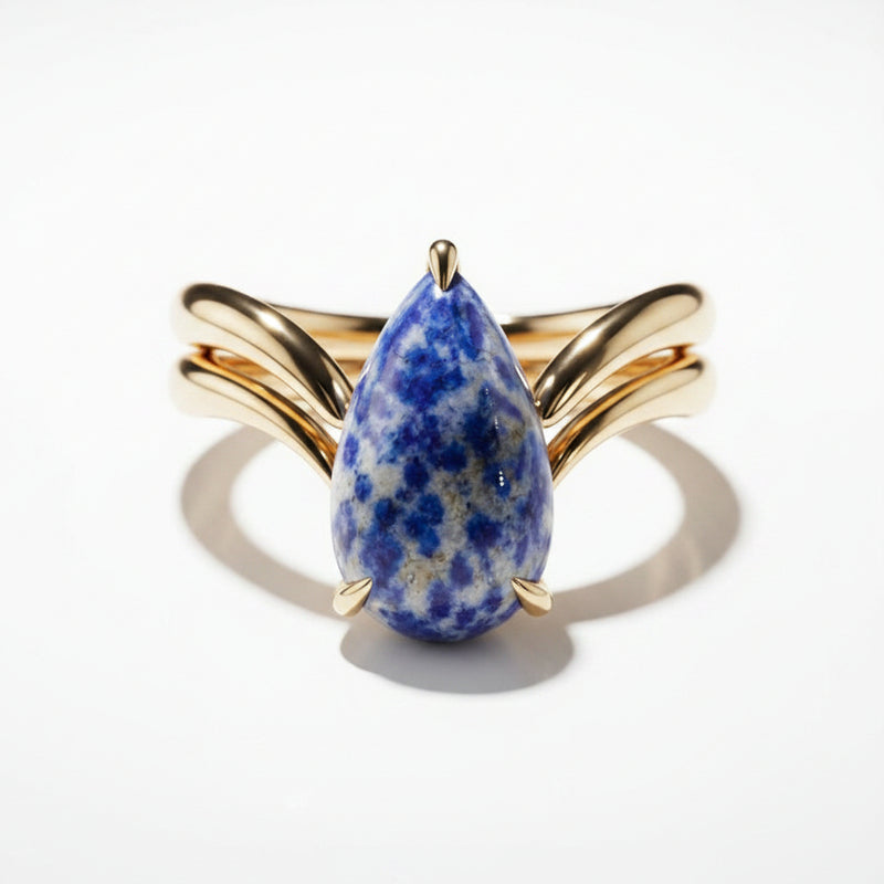 A gold ring featuring a teardrop-shaped blue and white spotted stone, displayed on a black marble surface.