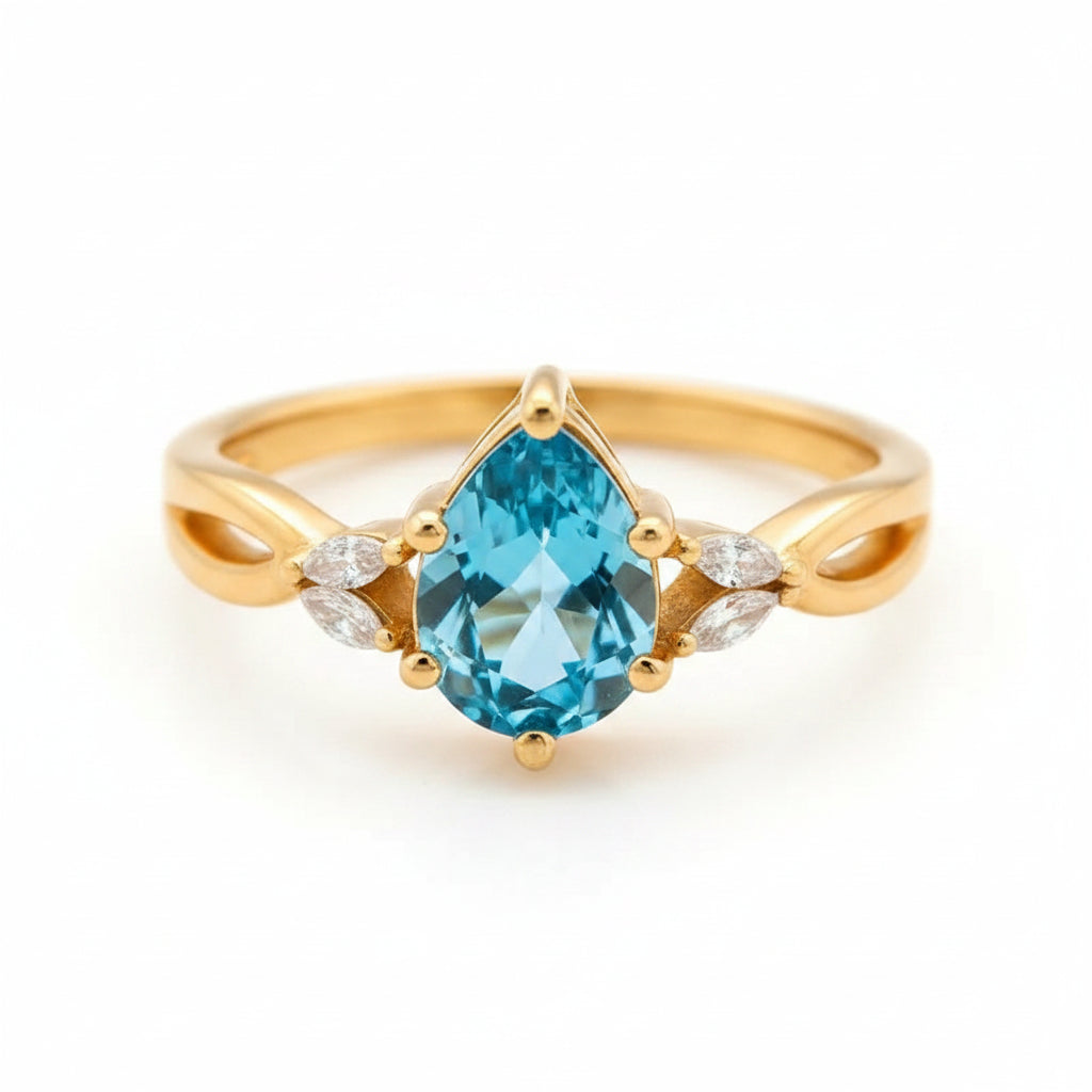A gold ring with a large teardrop-shaped blue gemstone and small clear accent stones on either side, photographed close up on a wooden surface with warm golden lighting.