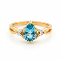 A gold ring with a large teardrop-shaped blue gemstone and small clear accent stones on either side, photographed close up on a wooden surface with warm golden lighting.
