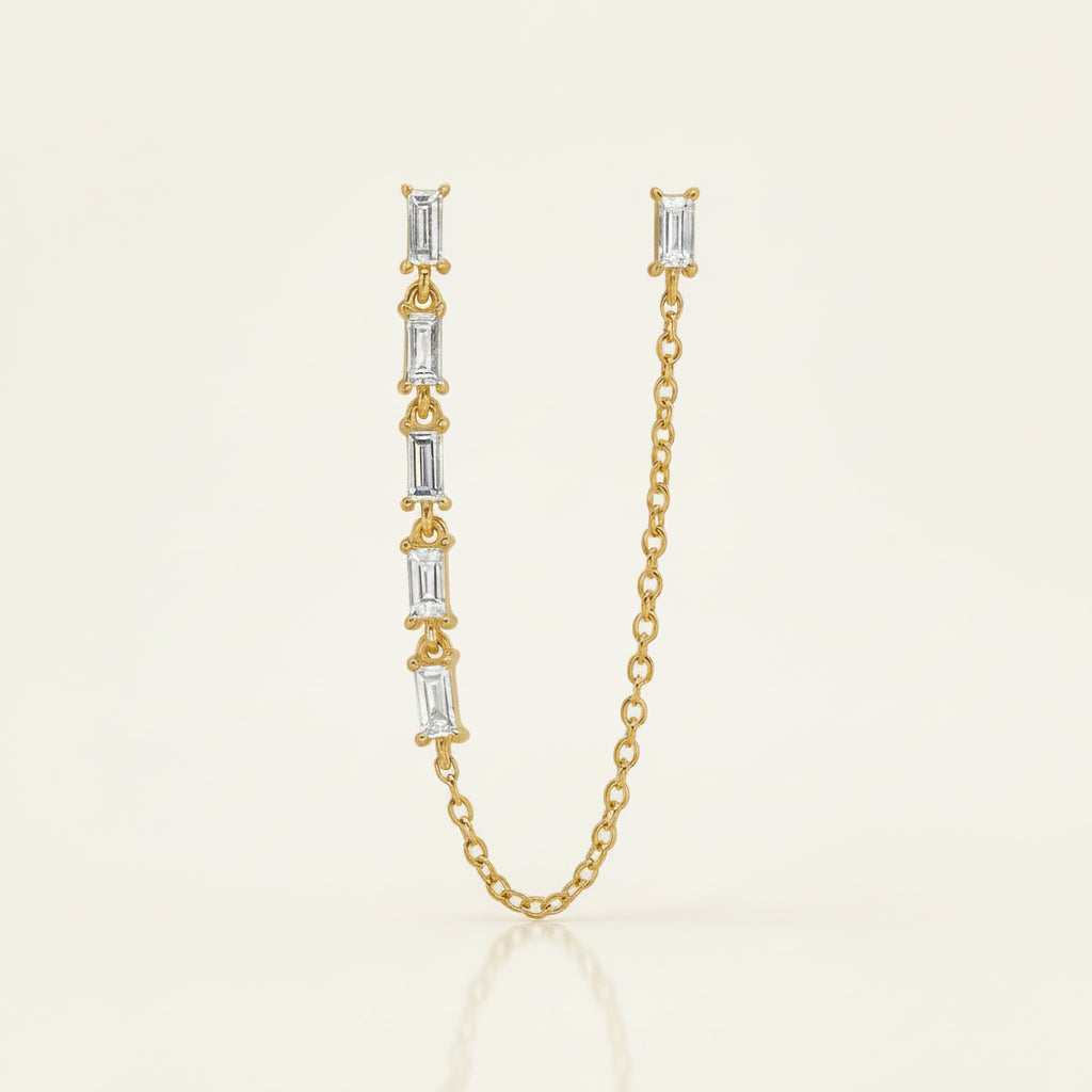 A gold chain necklace featuring a vertical bar with five baguette-cut clear stones in gold settings, shown on a white background with a faint diagonal grid pattern.