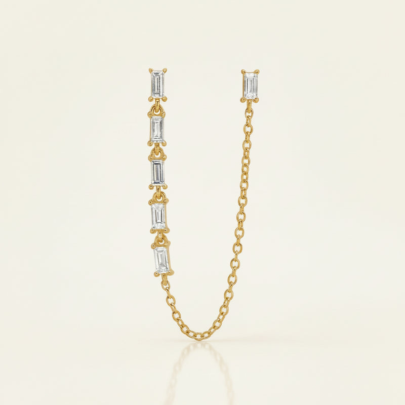 A gold chain necklace featuring a vertical bar with five baguette-cut clear stones in gold settings, shown on a white background with a faint diagonal grid pattern.