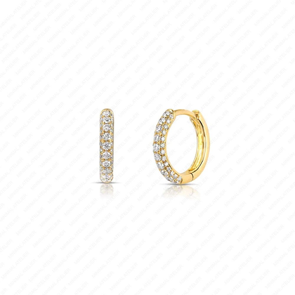 A pair of gold huggie hoop earrings adorned with pavé-set clear stones, shown from two angles on a white background with a faint diagonal grid pattern.