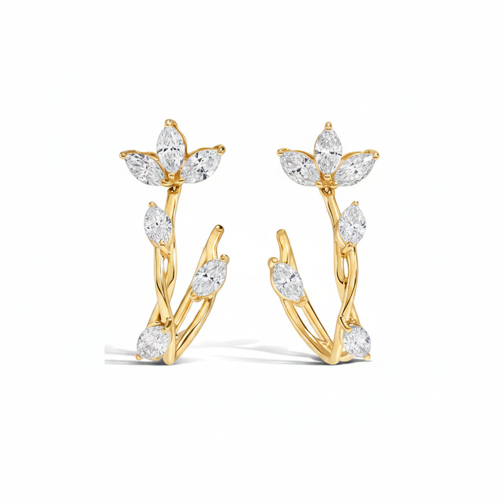 A pair of gold huggie hoop earrings decorated with marquise-cut clear gemstones arranged in a leafy branch design, shown on a white background with a faint diagonal watermark pattern.