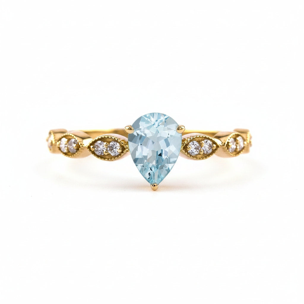 A yellow gold ring featuring a pear-cut light blue gemstone in the center, flanked by small oval and round clear accent stones on a white background with a faint watermark pattern.