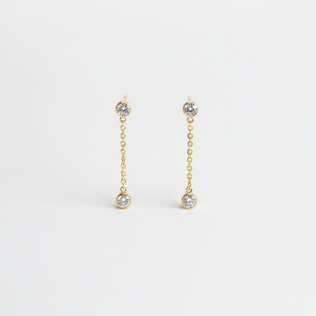 A pair of gold chain drop earrings with small clear gemstone accents, displayed on a light textured background.
