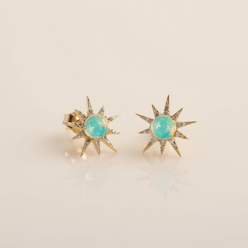 A pair of gold starburst stud earrings with oval light blue-green opal centers, displayed on a light gray background with a faint repeating watermark.