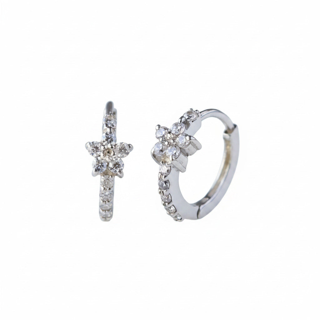 A pair of silver huggie hoop earrings decorated with small clear crystals, displayed on a light gray reflective surface.
