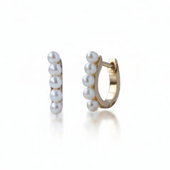 A pair of gold huggie hoop earrings decorated with five white freshwater pearls on each earring, shown against a white background with a faint diagonal watermark pattern.