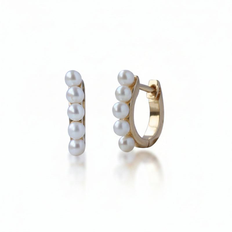 A pair of gold huggie hoop earrings decorated with five white freshwater pearls on each earring, shown against a white background with a faint diagonal watermark pattern.