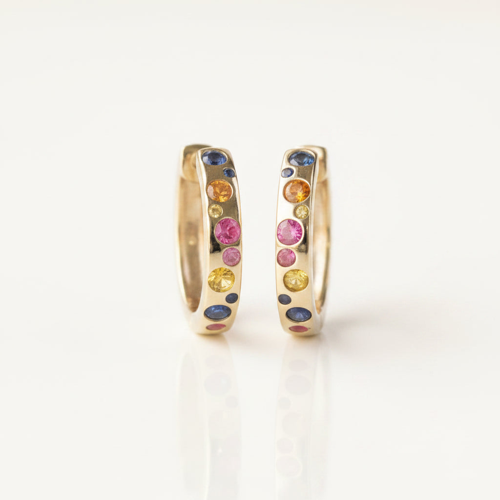 A pair of gold huggie hoop earrings with colorful gemstone accents, displayed on a white reflective surface with a repeating 'MINIMAL ATELIER' watermark background.