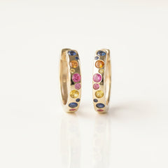 A pair of gold huggie hoop earrings with colorful gemstone accents, displayed on a white reflective surface with a repeating 'MINIMAL ATELIER' watermark background.
