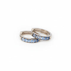 A pair of silver-toned huggie hoop rings with a row of light blue cubic zirconia stones, displayed on a textured white surface with a repeating 'MINIMAL ATTELIER' watermark pattern.