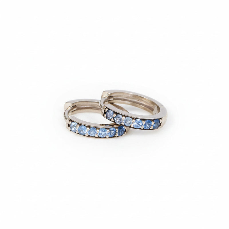 A pair of silver-toned huggie hoop rings with a row of light blue cubic zirconia stones, displayed on a textured white surface with a repeating 'MINIMAL ATTELIER' watermark pattern.