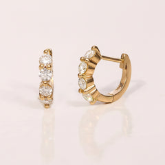A pair of gold huggie hoop earrings with clear cubic zirconia stones, shown from both front and side angles on a light pink background with a repeating 'MINIMAL ATELE' watermark.