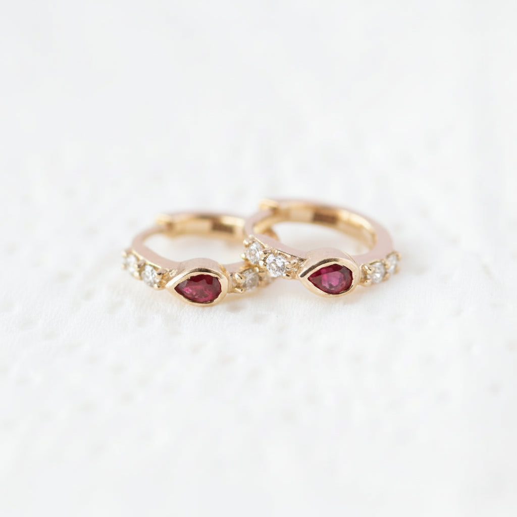 A pair of gold rings with teardrop-shaped red gemstones and small clear stones, displayed on a white textured surface with a diagonal 'MINIMAL ATELIER' watermark pattern.