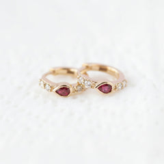 A pair of gold rings with teardrop-shaped red gemstones and small clear stones, displayed on a white textured surface with a diagonal 'MINIMAL ATELIER' watermark pattern.