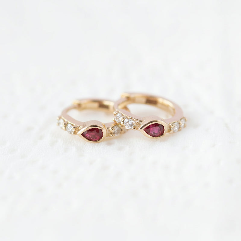 A pair of gold rings with teardrop-shaped red gemstones and small clear stones, displayed on a white textured surface with a diagonal 'MINIMAL ATELIER' watermark pattern.