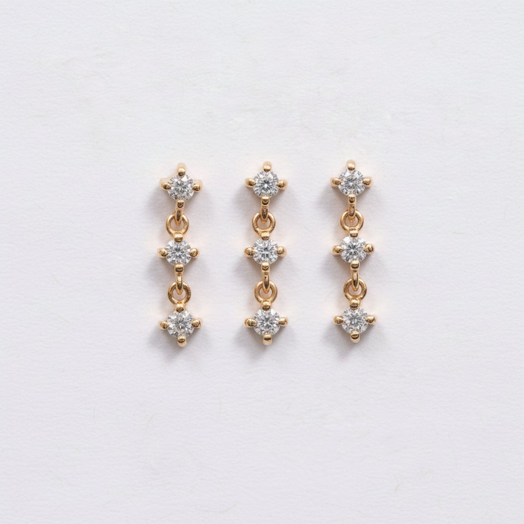 A pair of gold drop earrings with three clear round-cut stones in each earring, displayed on a light pink textured background with a repeating 'MINIMAL ATELIER' watermark.
