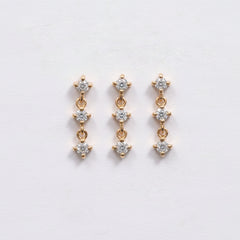 A pair of gold drop earrings with three clear round-cut stones in each earring, displayed on a light pink textured background with a repeating 'MINIMAL ATELIER' watermark.
