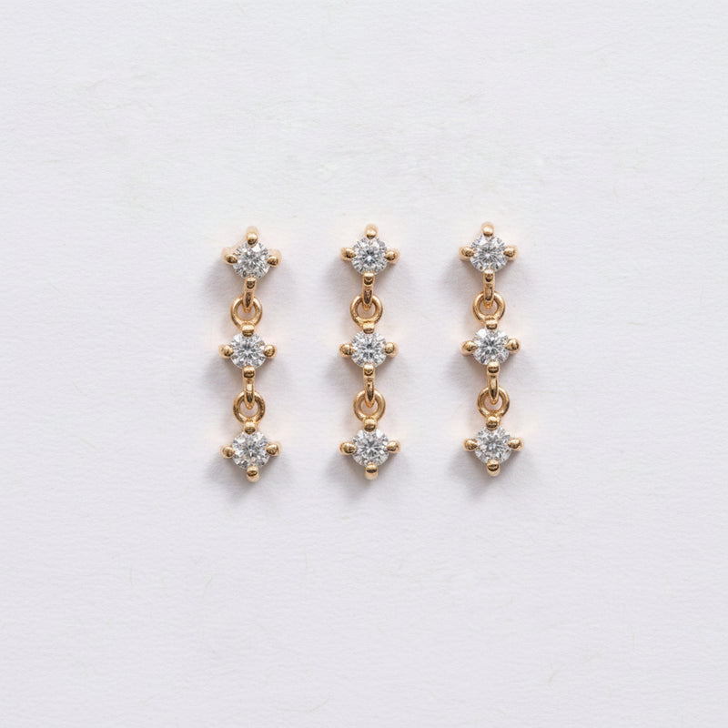 A pair of gold drop earrings with three clear round-cut stones in each earring, displayed on a light pink textured background with a repeating 'MINIMAL ATELIER' watermark.
