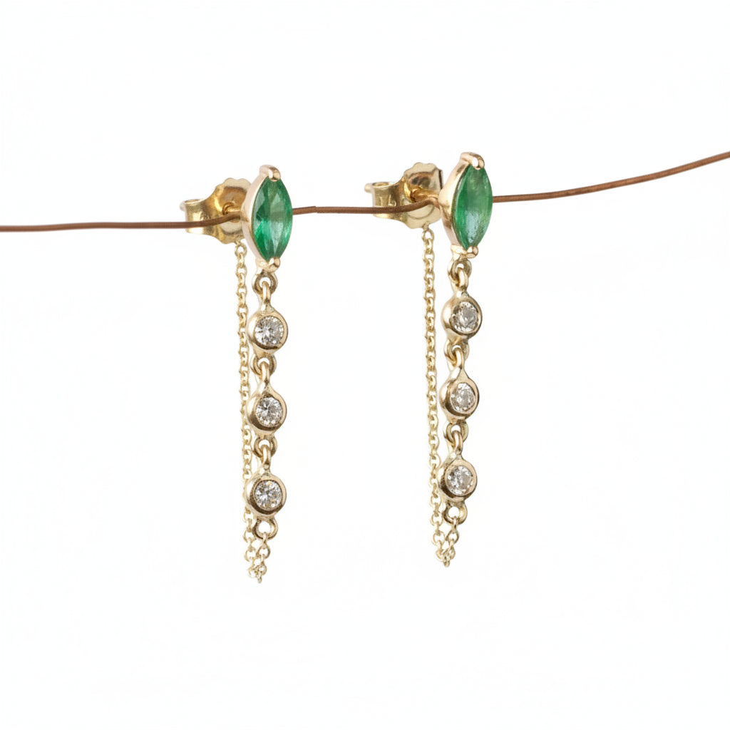 A pair of gold dangle earrings with marquise-cut green gemstones and clear round stones on a chain, displayed against a white background with a diagonal watermark pattern.