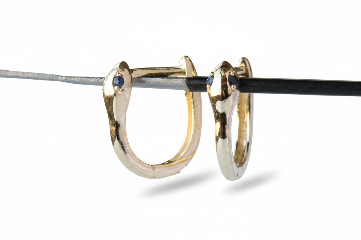 A pair of gold huggie hoop earrings with small blue gemstone accents on the front, shown hanging from a black rod against a gray background with a repeating watermark.