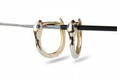A pair of gold huggie hoop earrings with small blue gemstone accents on the front, shown hanging from a black rod against a gray background with a repeating watermark.