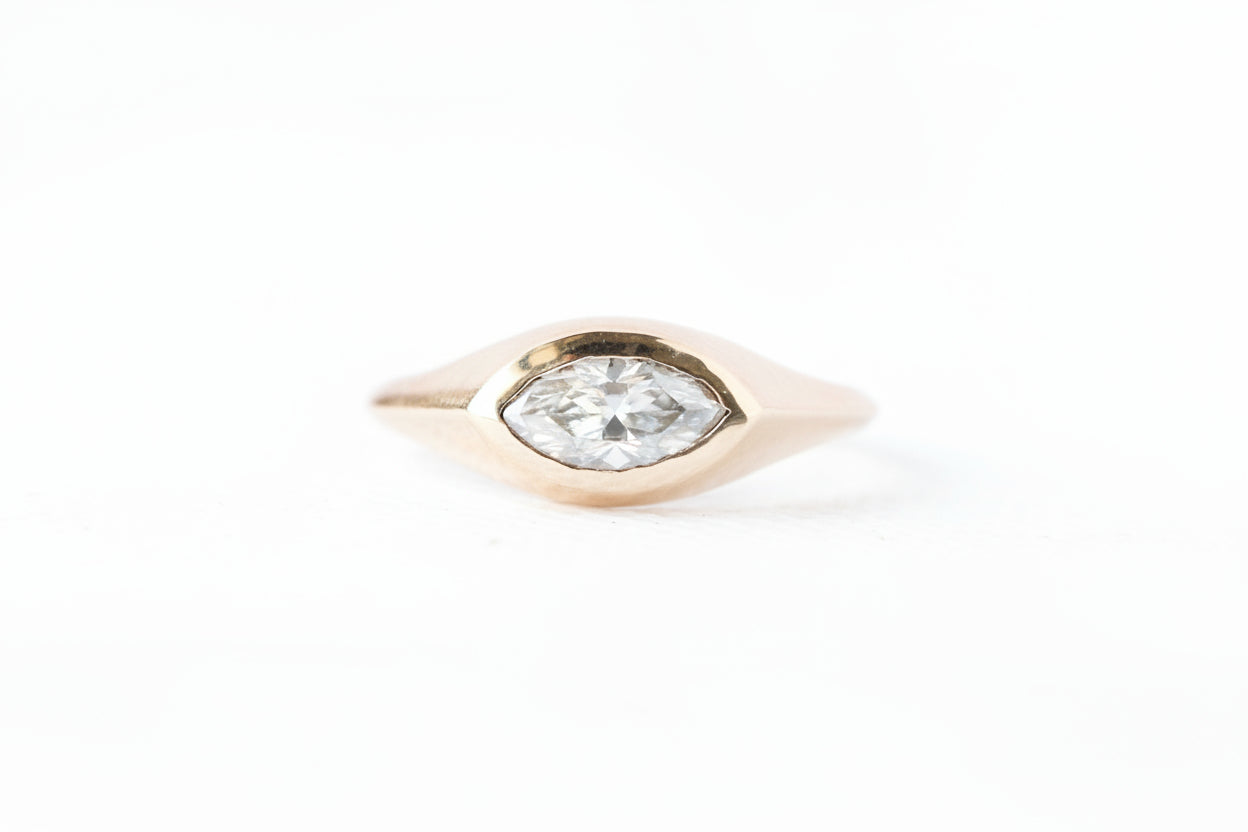 A gold ring featuring a marquise-cut clear gemstone in a marquise-shaped bezel setting, shown on a white background with a repeating 'MINIMAL ATELIER' watermark pattern.