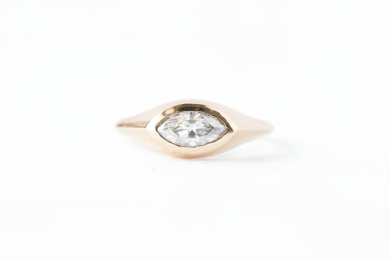A gold ring featuring a marquise-cut clear gemstone in a marquise-shaped bezel setting, shown on a white background with a repeating 'MINIMAL ATELIER' watermark pattern.