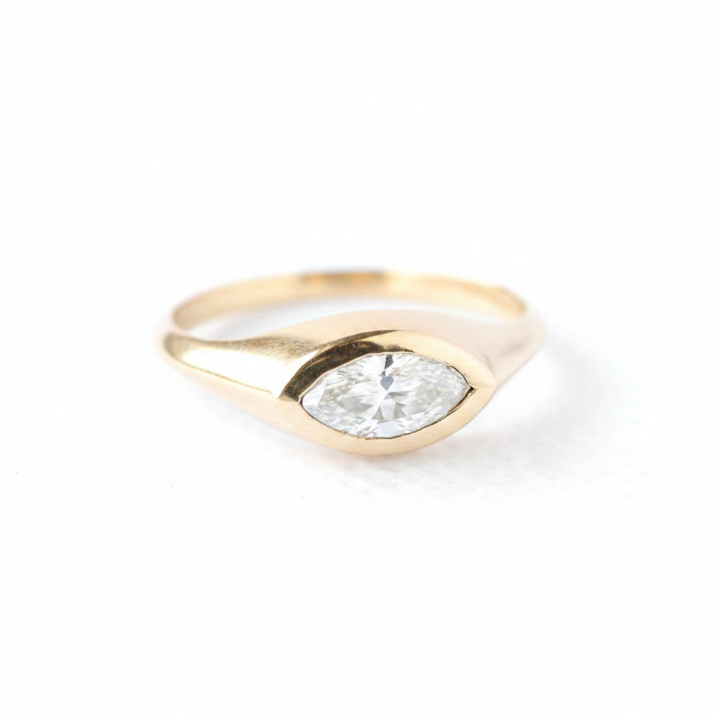 A gold ring with a marquise-cut clear gemstone, photographed on a white textured surface with a repeating 'MINIMAL ATELIER' watermark overlay.