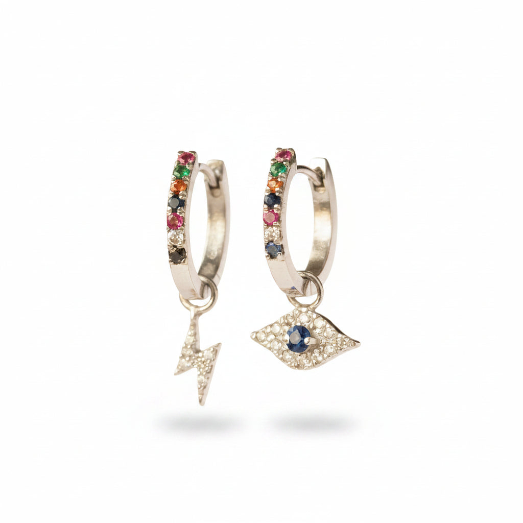 A pair of silver huggie hoop earrings with dangling charms, displayed on a metal stand. One earring features a vertical bar set with red, green, and black rhinestones, and the other features a dangling silver eye charm with a blue stone in the center.