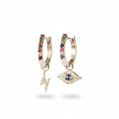 A pair of silver huggie hoop earrings with dangling charms, displayed on a metal stand. One earring features a vertical bar set with red, green, and black rhinestones, and the other features a dangling silver eye charm with a blue stone in the center.