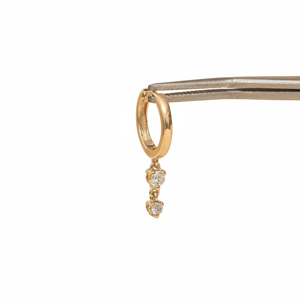 A small gold huggie hoop earring with two dangling clear gemstones, held by metal tweezers against a warm beige background with a repeating MINIMAL ATELIER watermark.