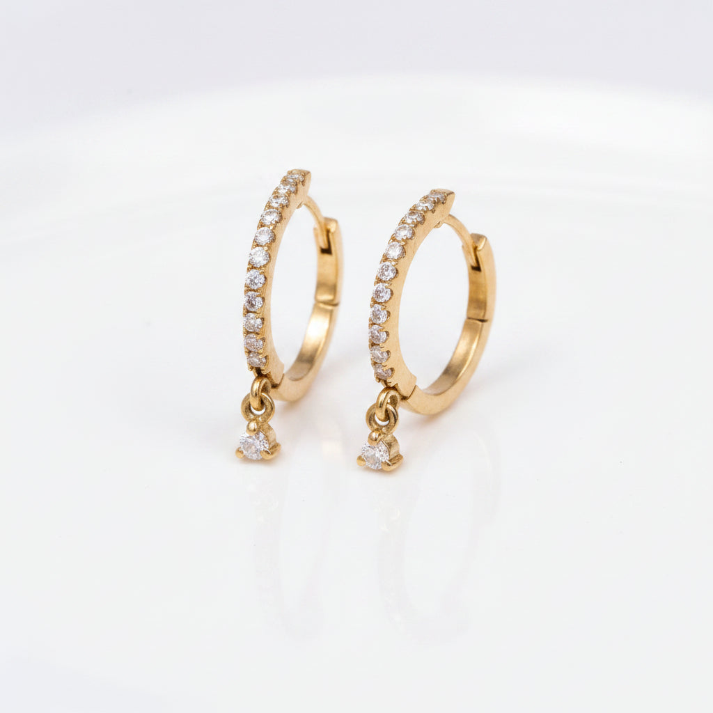 A pair of gold huggie hoop earrings adorned with clear pavé stones and dangling clear cubic zirconia drops, displayed on a soft beige textured fabric background.