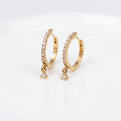 A pair of gold huggie hoop earrings adorned with clear pavé stones and dangling clear cubic zirconia drops, displayed on a soft beige textured fabric background.