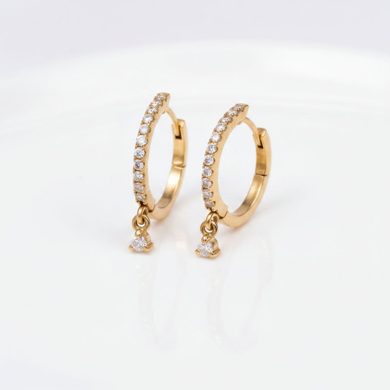 A pair of gold huggie hoop earrings adorned with clear pavé stones and dangling clear cubic zirconia drops, displayed on a soft beige textured fabric background.