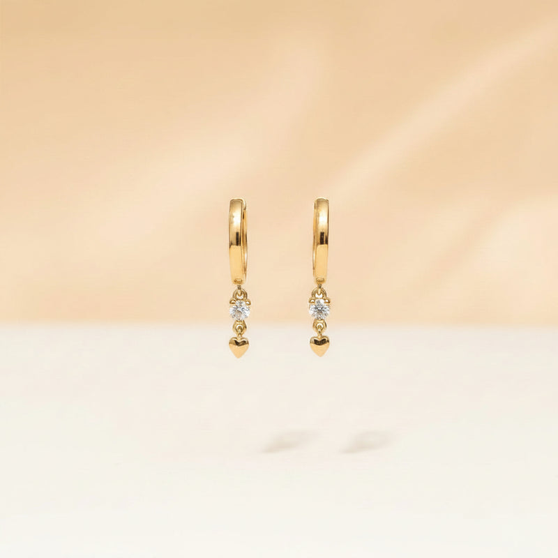 A close-up of a hand with lavender nail polish holding a pair of small gold hoop earrings with dangling clear gemstone drops against a warm beige background with a repeating 'MINIMAL ATELIER' watermark.