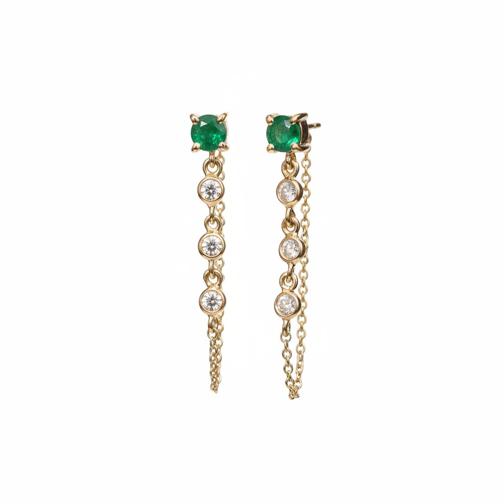 A pair of gold dangle earrings with round green emerald-cut stones at the top and a vertical line of clear cubic zirconia stones leading to a long chain, displayed on a beige and white textured background.